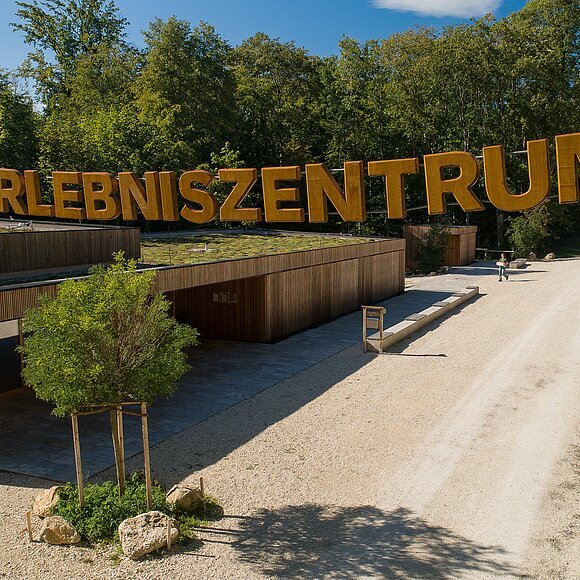 Walderlebniszentrum Schernfeld Am Walderlebniszentrum wird man am Eingang mit dem großen Schriftzug aus Holz empfangen. Darunter sieht man eine Holzhütte und rechts geht ein Feldweg in den Wald.