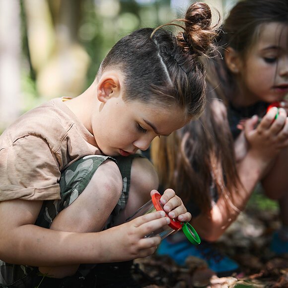 Kleine Forscher am WEZ Zwei Kinder sitzen am Waldboden und halten sehr interessiert etwas fest, dass sie zudem gebahnt anschauen.