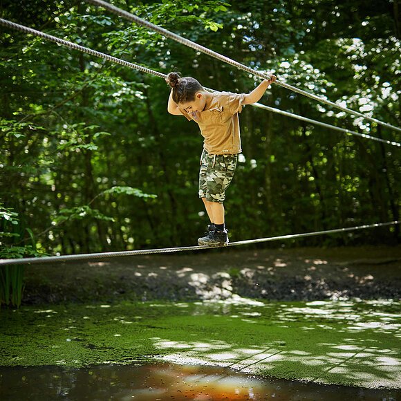 Kind auf dem Parcours am WEZ Ein Kind in gelbem Pulli überquert auf einem Seil mit Seilen zum Festhalten über dem Kopf einen Tülpel. Der Wald ist dunkgelgrün und hier und da scheint die Sonne durch.
