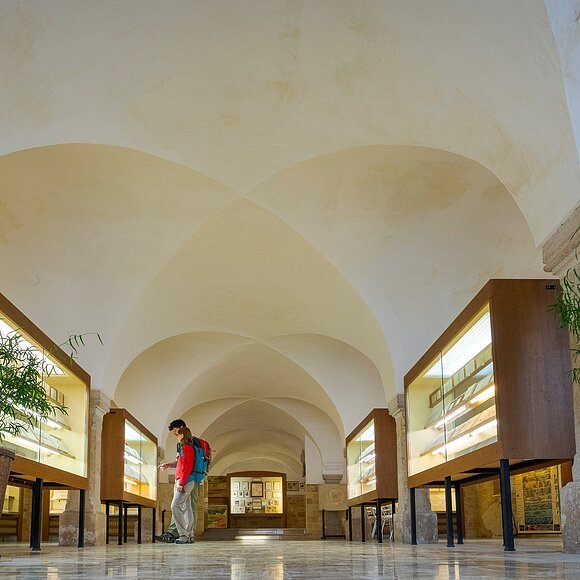 Im Mittelgang des Museums stehen links und rechts beleuchtete Vitrinen. Zwei Wanderer betrachten eine Vitrine auf der linken Seite. Der Raum ist hell und hat zudem Säulen und Rundbögen.