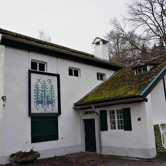 Weißes zweistöckiges Haus mit grünem Fensterladen und Moos auf dem Dach, im Herbstwald gelegen.