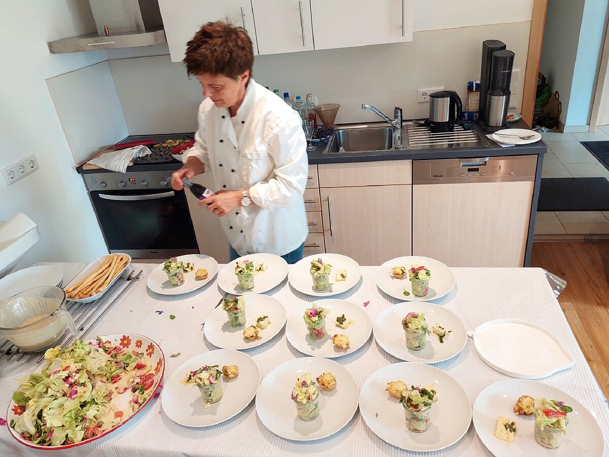 Brigitte Zinsmeister von Kräutertreff bei der Vorbereitung eines Kräutermenüs Eine Frau mit kurzen schwarzen Haare und in einer Kochjacke steht vor einem großen Tisch. Auf dem Tisch sind einige weiße Teller aufgereiht, welche mit einem Weckglas voll Salat, etwas Selbstgebackenem und Schafskäse gefüllt sind. Daneben ist eine große Schüssel mit buntem Salat, eine Schüssel mit einem weißen cremigen Dressing und eine Schale mit ein paar hellen Grissini zu sehen.